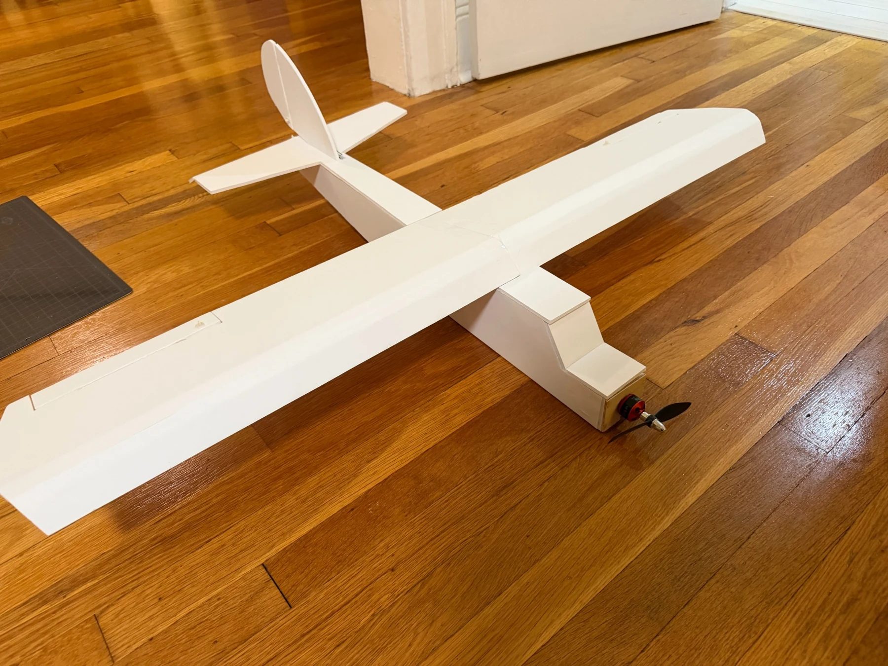 Remote-controlled foam board aircraft with motor, propeller, and tail assembly, photographed on a wooden floor.
